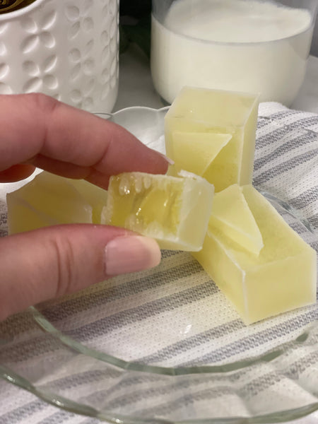 MonkGems are a sugar free crystal gummy candy. Lemon flavor crystal broken in half to show gummy inside and held in hand for scale against light background comprised of a white jar and heavy cream in a glass in the background on a gray striped towel.