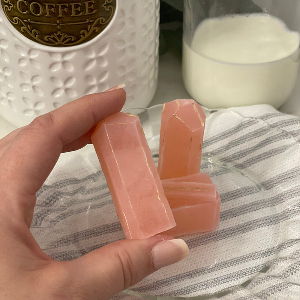 MonkGems are a sugar free crystal gummy candy. Raspberry flavor crystal being held in hand for scale against light background comprised of a white coffee jar and heavy cream in a glass in the background on a gray striped towel.